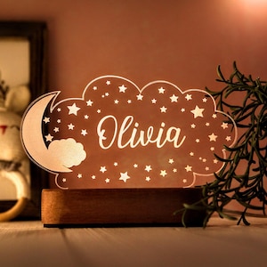 May include: A personalized night light featuring the name "Olivia" in elegant script. The design includes a cloud shape with stars and a crescent moon, set on a wooden base. The light emits a warm glow.
