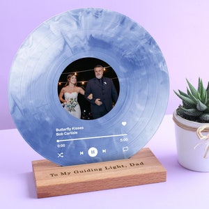 Puede incluir: Un disco de vinilo azul con una foto de una pareja en el centro. El disco está en un soporte de madera con el texto "To My Guiding Light, Dad" grabado en él. La canción "Butterfly Kisses" de Bob Carlisle está sonando en el disco.