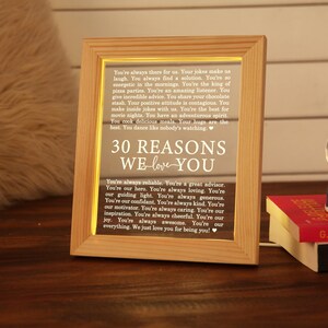 May include: A wooden frame with a light-up sign inside. The sign reads "30 Reasons We Love You" and lists reasons why someone is loved. The frame is sitting on a wooden table with a snow globe and a book.