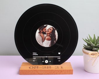 Könnte beinhalten: Eine schwarze Schallplatte mit einem Foto eines Paares in der Mitte. Die Schallplatte steht auf einem Holzständer mit der eingravierten Jahreszahl "08-28-21". Die Schallplatte hat eine Musikplayer-Oberfläche mit dem Titel "Yellow" von Coldplay, der abgespielt wird.