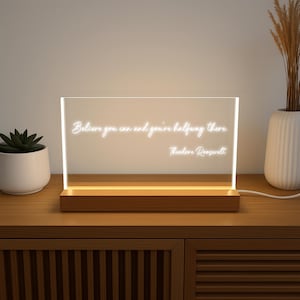 May include: A rectangular LED light with the quote "Believe you can and you're halfway there. Theodore Roosevelt" in white script. The light sits on a wooden base and is plugged into a wall outlet. A small succulent and vase are in the background.