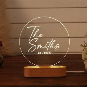 May include: A round, clear acrylic sign with a wooden base. The sign has the text "The Smiths EST. 8/6/20" in white script.