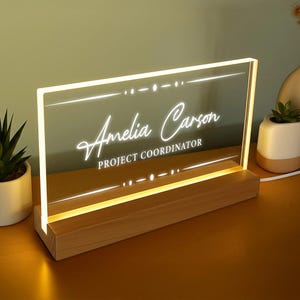 May include: A personalised desk nameplate with the name "Amelia Carson" and the title "PROJECT COORDINATOR" illuminated in white script. The clear acrylic panel is set in a wooden base, emitting a warm glow. Decorative lines and dots frame the text.