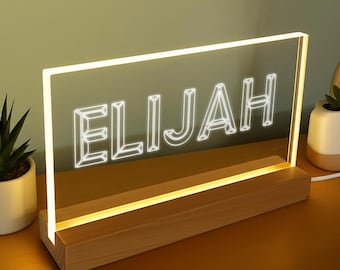 May include: A lighted sign with the name "ELIJAH" in white, outlined letters. The sign is rectangular and sits on a wooden base. The light glows around the edges and the letters. The background is a soft green.
