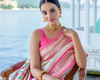 May include: A woman wearing a pink top and a traditional Indian sari with gold jewelry. The sari features a cream-colored base with pink and gold accents. The jewelry includes a necklace, earrings, a ring, and a bracelet.