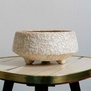 May include: A low, round ceramic planter featuring a textured, off-white glaze with a speckled, sandy appearance. The bowl-shaped vessel is supported by three small, integrated feet and has a wide, open top with a visible terracotta-coloured rim.