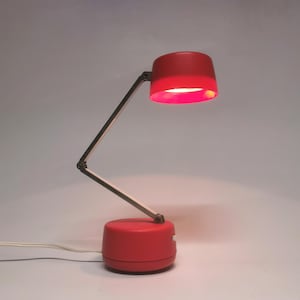 Könnte beinhalten: Rote Schreibtischlampe mit zylindrischem Sockel und passendem Lampenschirm. Der schwarze und silberne Gelenkarm ermöglicht eine flexible Positionierung. Ein weißes Netzkabel ist angeschlossen. Modernes Design.