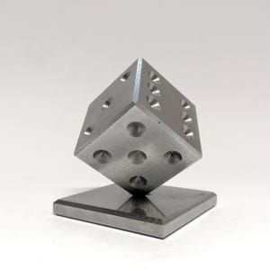 Puede incluir: Una escultura de dado metálico grande con hendiduras circulares perforadas que representan los números del 1 al 6. El dado está equilibrado sobre una esquina encima de una base cuadrada, todo en color gris plateado. Un artículo decorativo único.