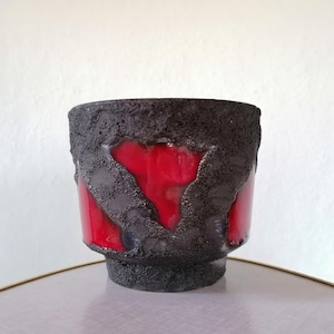 Op de afbeelding: Een cilindrische keramische plantenpot met een ruwe, donker antracietkleurige textuur aan de buitenkant die contrasteert met gladde, glanzende, felrode geglazuurde delen die zichtbaar zijn door de onregelmatige openingen in de laag.