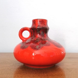 Könnte beinhalten: Eine leuchtend rote Keramikvase mit rundem Körper und kleinem Henkel. Die Vase hat eine glänzende Oberfläche mit dunkelbraunen, gesprenkelten Details am Hals. Das Design ist ein klassischer, moderner Stil aus der Mitte des Jahrhunderts, ideal für die Inneneinrichtung.