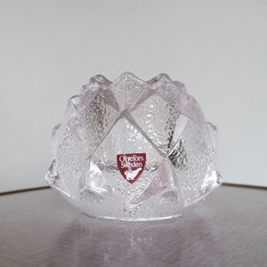 Könnte beinhalten: Ein durchsichtiger Kerzenhalter aus Glas mit geometrischem Design. Der Halter hat eine strukturierte Oberfläche und trägt ein rotes Etikett mit dem Text "Orrefors Sweden".