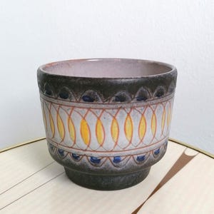 May include: A ceramic planter with a cylindrical shape, featuring a dark brown base and rim. The center band displays a repeating pattern of orange and yellow oval shapes outlined in thin lines, accented by small blue dots within dark scalloped arches.