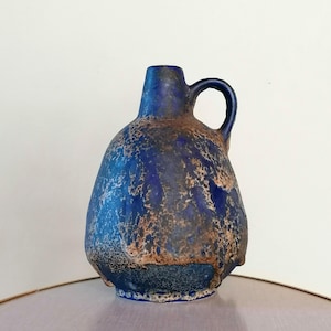 May include: A ceramic pitcher with a rounded body and a single handle. The surface features a textured, mottled glaze in deep cobalt blue and earthy brown tones, creating a rugged, uneven appearance across the entire exterior of the vessel.