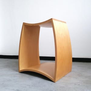 Könnte beinhalten: Ein hellfarbener Holzstuhl mit geschwungenem Design. Der Hocker hat eine rechteckige Basis und eine geschwungene Oberseite mit offenen Seiten. Das Holz hat eine natürliche Maserung und eine glatte Oberfläche. Der Hocker ist etwa 46 cm hoch.