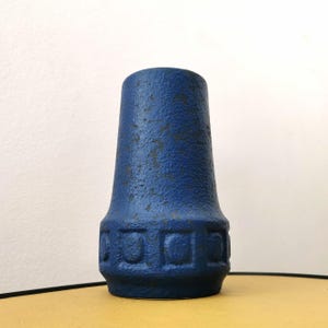 Könnte beinhalten: Eine blaue Keramikvase mit strukturierter Oberfläche und geometrischem Muster am Boden. Die Vase hat einen ausgestellten oberen Teil und eine dunkelblaue Farbe mit einigen Abnutzungsstellen.