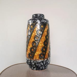Peut inclure: Un vase en céramique cylindrique avec un motif noir, orange et blanc. Le vase présente un motif répété de cercles noirs et de rayures diagonales. La base et le bord supérieur sont noirs avec des mouchetures blanches, tandis que le centre a des tons orange et jaune.
