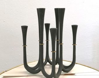 以下が含まれることがあります： 7つのキャンドルホルダーが付いた、濃いグレーの金属製燭台。 ホルダーは曲線的で対称的なデザインで配置され、各ホルダーの基部には真鍮色のリングが付いています。 燭台は模様のある表面にあります。