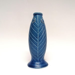 Könnte beinhalten: Eine blaue Keramikvase mit einem fischähnlichen Design. Die Vase hat eine strukturierte Oberfläche mit einer Mittellinie und diagonalen Linien, die einem Blatt ähneln. Sie hat zwei kreisförmige, augenähnliche Details in der Nähe der Oberseite und ist etwa 25 cm hoch.