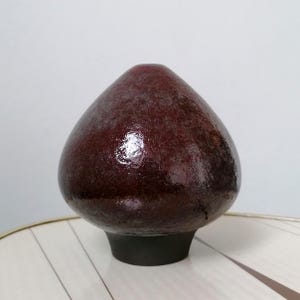 Può includere: Un vaso in ceramica color bordeaux con una forma arrotondata a goccia. Il vaso ha una superficie strutturata e poggia su una base corta e nera. La finitura lucida riflette la luce.
