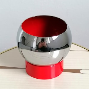 May include: A modern, spherical decorative bowl with a polished silver exterior and a vibrant red interior. The bowl sits on a matching red base, creating a striking contrast. The design is sleek and contemporary.