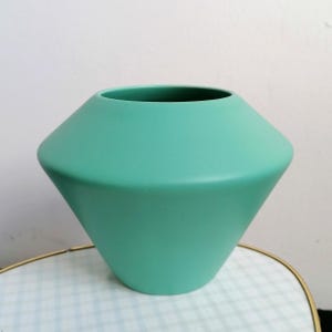 Könnte beinhalten: Eine matte, mintgrüne Keramikvase mit einer breiten, kantigen geometrischen Silhouette. Das Gefäß hat eine breite, abfallende Schulter, die sich zu einer schmaleren Basis hin verjüngt, mit einer runden Öffnung oben und einer glatten Oberfläche.