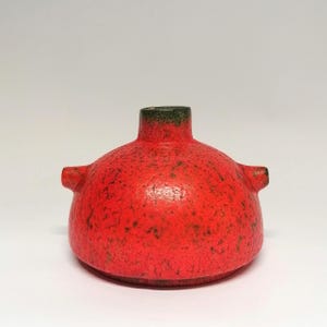 Könnte beinhalten: Eine leuchtend rote Keramikvase mit einem runden, bauchigen Körper und einem kurzen, schmalen Hals. Die Oberfläche weist eine texturierte, rissige Optik mit dunkelgrünen, gesprenkelten Akzenten auf. Zwei kleine Griffe ragen seitlich hervor.
