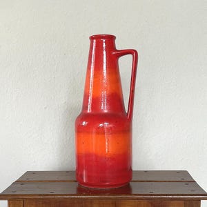 Op de afbeelding: Een hoge keramische vaas met een handvat, met een verloop van rode en oranje tinten. De vaas heeft een cilindrische vorm die naar boven toe smaller wordt, met een handvat. Het staat op een houten oppervlak.