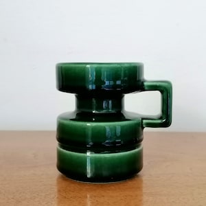 Könnte beinhalten: Eine dunkelgrüne Keramiktasse mit einem einzigartigen, geometrischen Design. Die Tasse hat einen zylindrischen Körper mit einem Griff und einer ausgestellten Oberseite. Die glänzende Oberfläche reflektiert das Licht und hebt die Form und Details der Tasse hervor. Die Tasse steht auf einer Holzoberfläche.