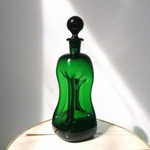 Könnte beinhalten: Eine hohe, smaragdgrüne Glasdekanter mit rundem Boden und einem kugelförmigen Verschluss. Der Dekanter hat eine einzigartige, geschwungene Form und einen langen, schlanken Hals. Der Verschluss ist eine dunkle, massive Kugel. Der Dekanter ist leer.