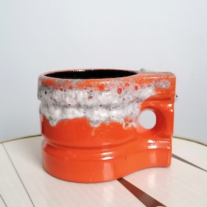 Könnte beinhalten: Leuchtend orangefarbene Keramiktasse mit schwarzem Innenraum und weißem, strukturiertem Band. Die Tasse hat ein einzigartiges, geometrisches Design mit Henkel und kreisförmiger Öffnung. Die Tasse steht auf einer gemusterten Oberfläche.