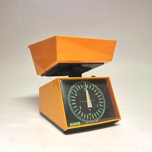 May include: A retro orange and black kitchen scale. The scale features a circular dial with green numerals and markings, and a white needle. The scale has a rectangular base and a tray. The dial reads in kilograms.