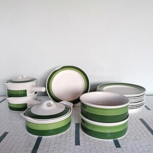 Op de afbeelding: Een verzameling wit keramisch servies met groene strepen. De set bevat borden, kommen en serveerschalen met deksels. De groene strepen zijn een opvallend accent op de verder witte stukken. Het servies staat op een patroon oppervlak.
