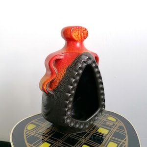 Könnte beinhalten: Eine Keramikvase mit einzigartigem Design. Die Oberseite ist leuchtend rot, geht in Orange und dann in Schwarz über. Die Vase hat eine große Öffnung und einen strukturierten, gepunkteten Rand. Sie steht auf einem gemusterten Tisch.