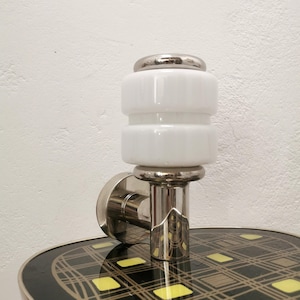 Puede incluir: Una lámpara de pared vintage con una pantalla de vidrio blanco apilado y una base cromada. La lámpara está montada sobre una mesa con un patrón geométrico negro y dorado. Un elemento decorativo elegante.