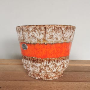 Puede incluir: Una maceta de cerámica con un exterior texturizado, moteado en marrón y blanco. Una vibrante banda naranja rodea el centro. La maceta tiene una forma ligeramente acampanada y una pequeña pegatina redonda en la parte delantera.