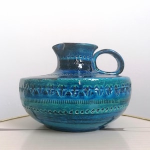 Może przedstawiać: Ceramiczny dzbanek w odcieniach niebieskiego i turkusowego z uchwytem. Dzbanek ma zaokrąglony korpus z teksturowanym, geometrycznym wzorem u podstawy i gładkie, błyszczące wykończenie. Otwór dzbanka jest rozszerzony.