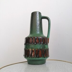 May include: A tall ceramic pitcher vase featuring a textured green finish with a dark brown band around the center. The surface has a rough, tactile quality with vertical brown stripes integrated into the body and a single handle on the side.