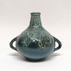 Könnte beinhalten: Eine handgefertigte Keramikvase mit rundem Körper und zwei kleinen Griffen. Die Vase hat einen Farbverlauf von blau-grünen Farbtönen, mit dunklerem Sockel und heller, strukturierter Glasur oben. Gesamthöhe ca. 15 cm.