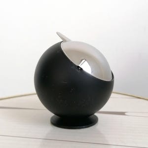 Può includere: Posacenere sferico nero opaco con coperchio argentato a forma di semicerchio. Il posacenere ha una base piccola e rotonda e una piccola linguetta appuntita sul coperchio.
