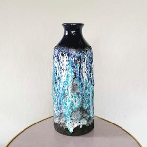 Könnte beinhalten: Eine hohe Keramikvase mit einem dunkelblauen, glänzenden Oberteil und einem strukturierten Körper. Der Körper weist ein fließendes Design aus weißen, türkisfarbenen und blauen Farbtönen über einem schwarzen Sockel auf. Die Vase hat einen schmalen Hals und eine rechteckige Form.