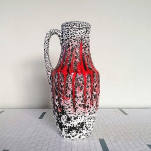 Könnte beinhalten: Eine hohe Keramikvase mit Henkel, verziert mit einem gesprenkelten Muster in Weiß und Schwarz. Der Mittelteil hat ein auffälliges rotes, tropfendes Design. Ein dekoratives Objekt.