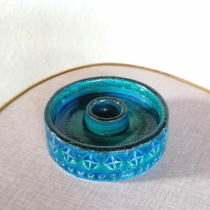 Può includere: Un portacandele in ceramica turchese e verde acqua. Il portacandele ha una forma circolare con un centro rialzato per la candela. L'esterno è decorato con un motivo geometrico e dettagli argentati. L'interno ha una finitura lucida.