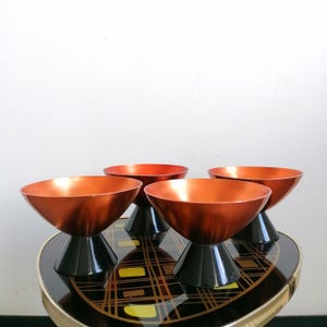 Könnte beinhalten: Vier kupferfarbene Schalen mit schwarzen Sockeln. Die Schalen haben ein breites, flaches Design und sind auf einer gemusterten Oberfläche angeordnet. Das Innere der Schalen reflektiert das Licht und erzeugt einen warmen Schein.