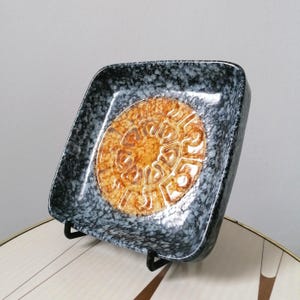 May include: A square ceramic dish featuring a black and white speckled glaze pattern. The center of the dish displays a circular, embossed golden-brown design with a series of repeating, curved, and geometric shapes arranged in a radial pattern.