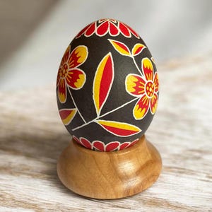 Op de afbeelding: Een donkerbruin ei versierd met handgeschilderde rode en gele bloemmotieven met bloemblaadjes en bladeren, rustend op een gladde, lichtgekleurde houten standaard. Het ontwerp bedekt het oppervlak van het ei in een symmetrische opstelling.