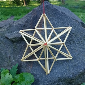 Peut inclure: Une étoile à six branches fabriquée à partir de tiges de paille beige clair. La structure géométrique présente de multiples lignes entrecroisées formant une étoile complexe en trois dimensions, maintenue par un fin fil rouge au centre.