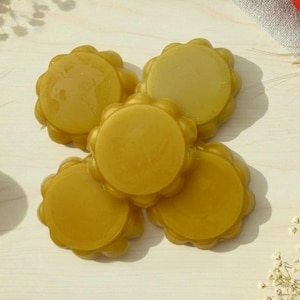 Puede incluir: Vista aérea de cinco jabones dorados en forma de flor dispuestos sobre una superficie de madera clara. Se ven huevos de Pascua decorativos azules y rojos con diseños intrincados, junto con ramitas de flores blancas.