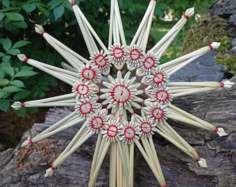 May include: A straw star with a red and white center, made of woven straw with red string accents. The star is a traditional Christmas decoration.
