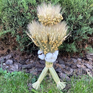 Peut inclure: Une gerbe décorative composée de tiges de blé séchées et de petites fleurs de lin jaunes, agrémentée de boules de coton blanc. Les tiges sont assemblées à la base avec de la paille tressée, formant une structure en trépied à deux pieds.