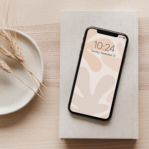 K&ouml;nnte beinhalten: Ein schwarzes Smartphone mit einer abstrakten Tapete in Wei&szlig; und Beige. Die Uhrzeit ist 10:24 Uhr und das Datum ist Dienstag, 13. September.